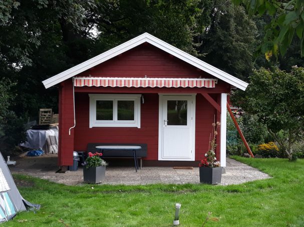 Rotes Gartenhaus mit weißem Tür und Fenster, umgeben von Grünflächen und Pflanzen.