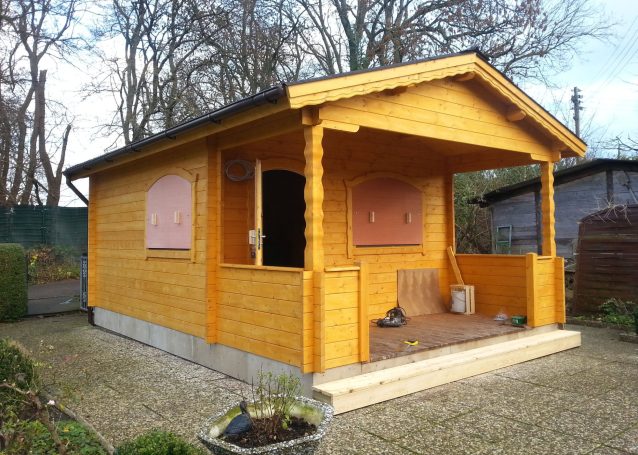 Holzhütte mit Veranda, leicht erhöht, umgeben von Gartenfläche und Bäumen.